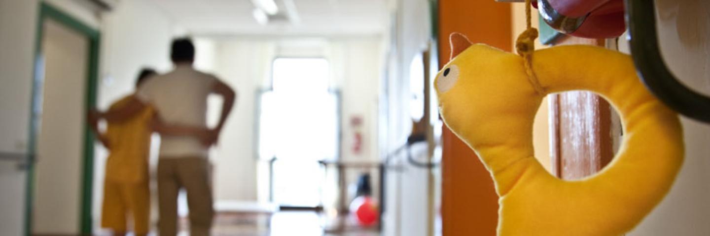 Un ragazzino sordocieco cammina con il supporto di un adulto per i corridoi del Centro di Osimo. I due sono in secondo piano, mentre in primo piano è messo a fuoco il dettaglio di uno dei segnalini oggettuali che identificano le porte dei vari ambienti del Centro