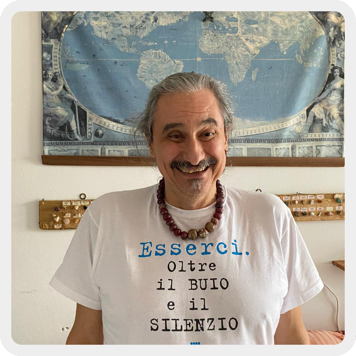 Roberto, uomo sordocieco di circa 60 anni, sorride indossando una t-shirt in cui c'è scritto "Esserci oltre il buio e il silenzio". Indossa anche una collana di legno. Sullo sfondo una mappa del mondo ed alcuni minerali. 