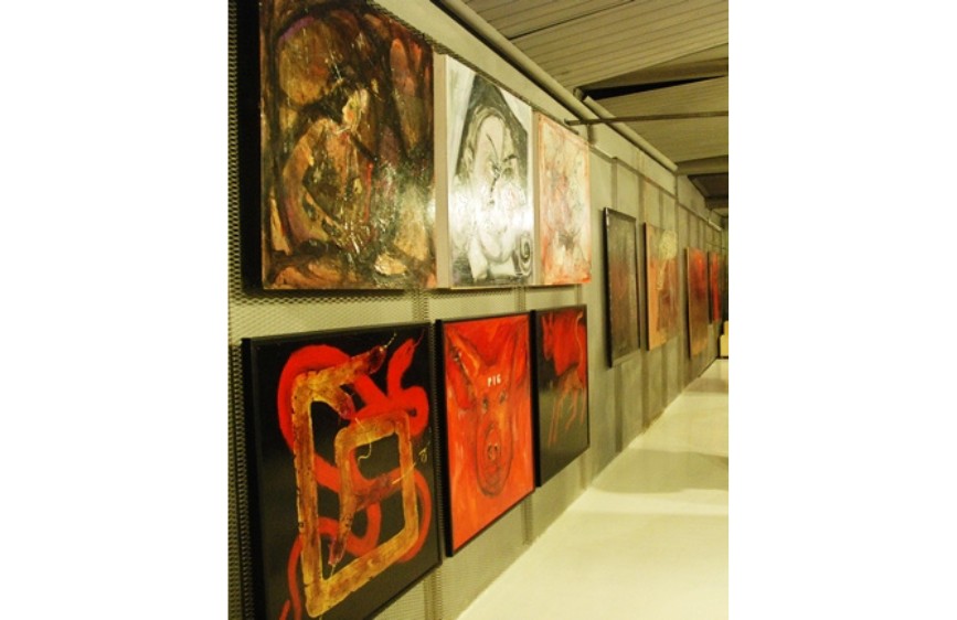 L'immagine mostra una galleria d'arte moderna con due file di quadri appesi lungo una parete grigia, disposti in prospettiva. I dipinti in primo piano presentano colori vividi, in particolare rosso e nero, mentre quelli più in lontananza hanno tonalità più scure e variegate.