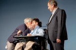 Antonio è seduto sulla sedia a rotelle. Viene baciato dalla mamma che è alla sua destra, mentre il papà osserva felice al suo fianco sinistro in piedi