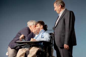 Antonio è seduto sulla sedia a rotelle. Viene baciato dalla mamma che è alla sua destra, mentre il papà osserva felice al suo fianco sinistro in piedi