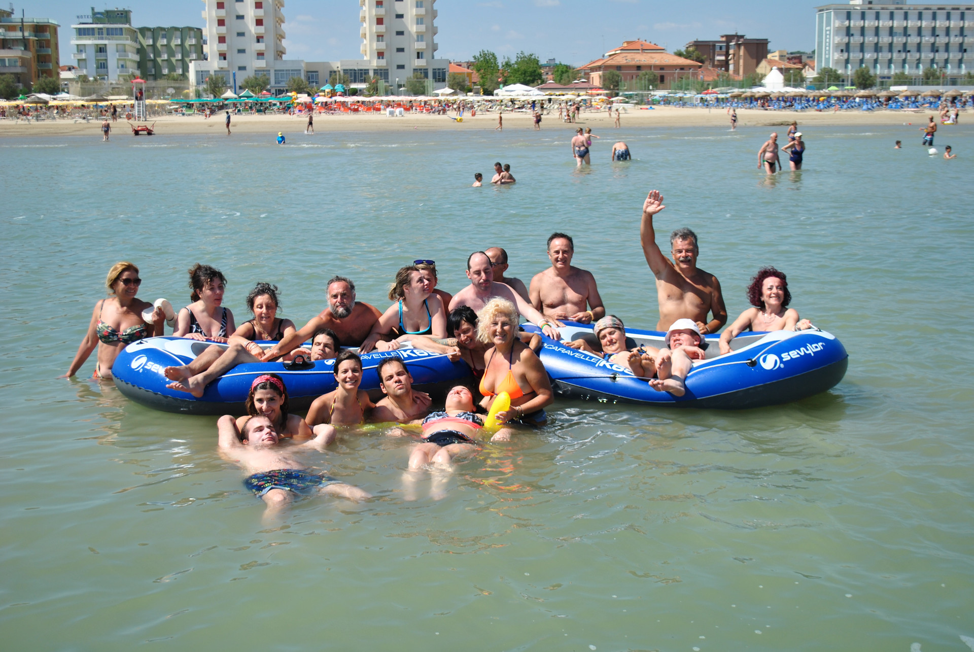 Foto di gruppo al mare durante un soggiorno estivo a Senigallia. Volontari, educatori e utenti fanno il bagno insieme, raggruppati intorno e a bordo di due gommoni gonfiabili, e salutano verso chi sta scattando la foto