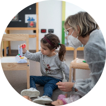 Letizia, bambina con la sindrome di Charge, gioca con la sua educatrice, Manola. Letizia è seduta per terra e inserisce le ciambelline nel pallottoliere divise per colore. Foto scattata durante il periodo del Covid perchè Manola indossa camice e mascherina.