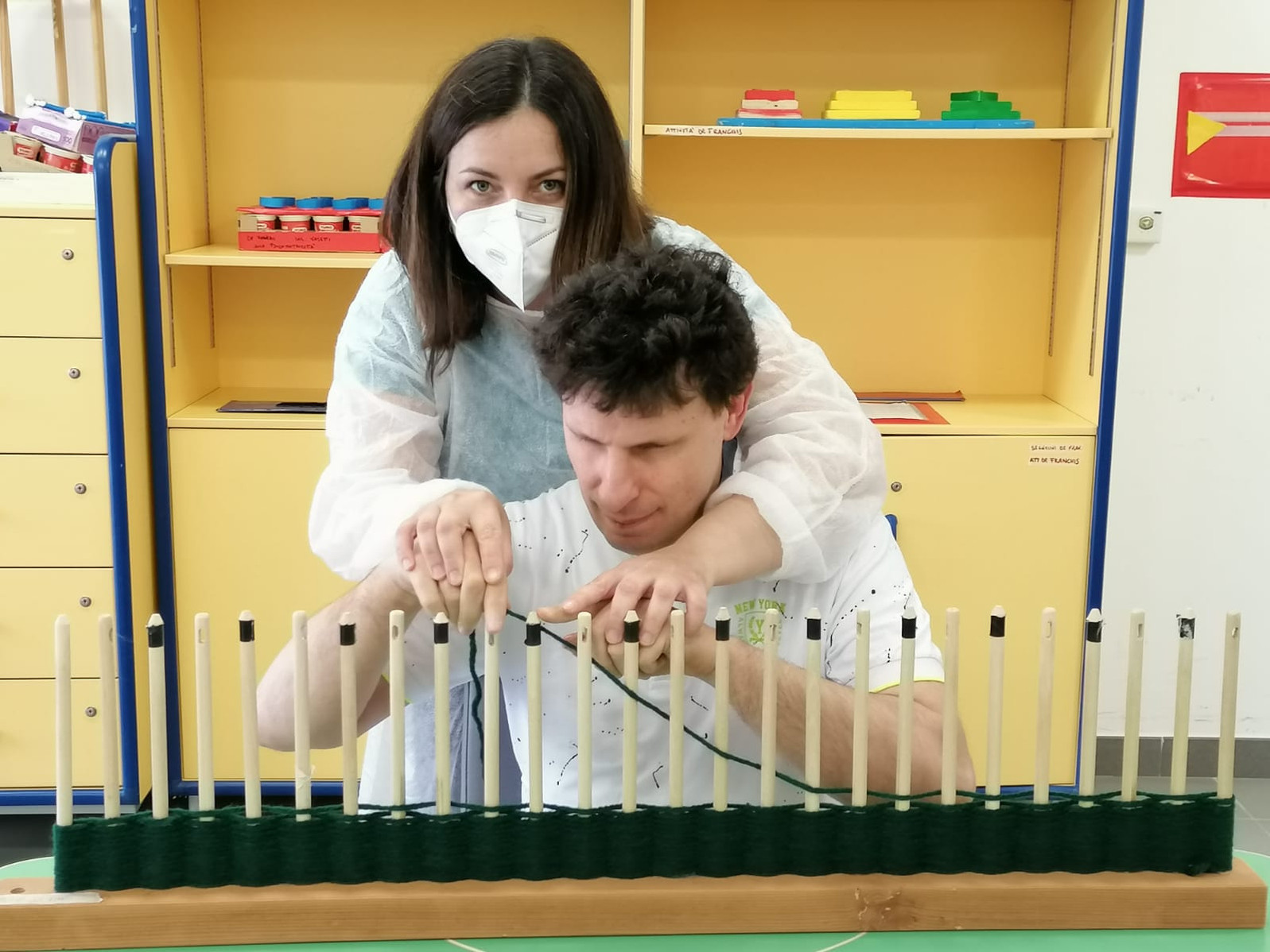 Francesca l'educatrice con indosso mascherina e camice monouso guida i gesti di un utente adulto che lavora nel laboratorio tessile del Centro della Lega del Filo d'Oro a Termini Imerese (PA)