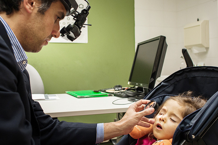 Il Dr.Pantanetti visita una piccola utente nell'ambulatorio oculistico di Osimo. La bambina è seduta nel passeggino, mentre l'oculistale osserva l'occhio destro con una lente d'ingrandimento.