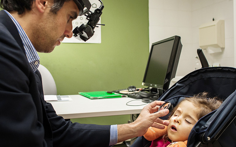 Il Dr.Pantanetti visita una piccola utente nell'ambulatorio oculistico di Osimo. La bambina è seduta nel passeggino, mentre l'oculistale osserva l'occhio destro con una lente d'ingrandimento.