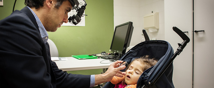 Il Dr.Pantanetti visita una piccola utente nell'ambulatorio oculistico di Osimo. La bambina è seduta nel passeggino, mentre l'oculista le osserva l'occhio destro con una lente d'ingrandimento.
