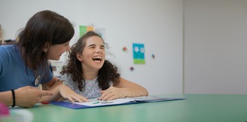 Giulia, bambina utente della Lega del Filo d’Oro, sorride mentre legge un libro tattile insieme a un’operatrice.