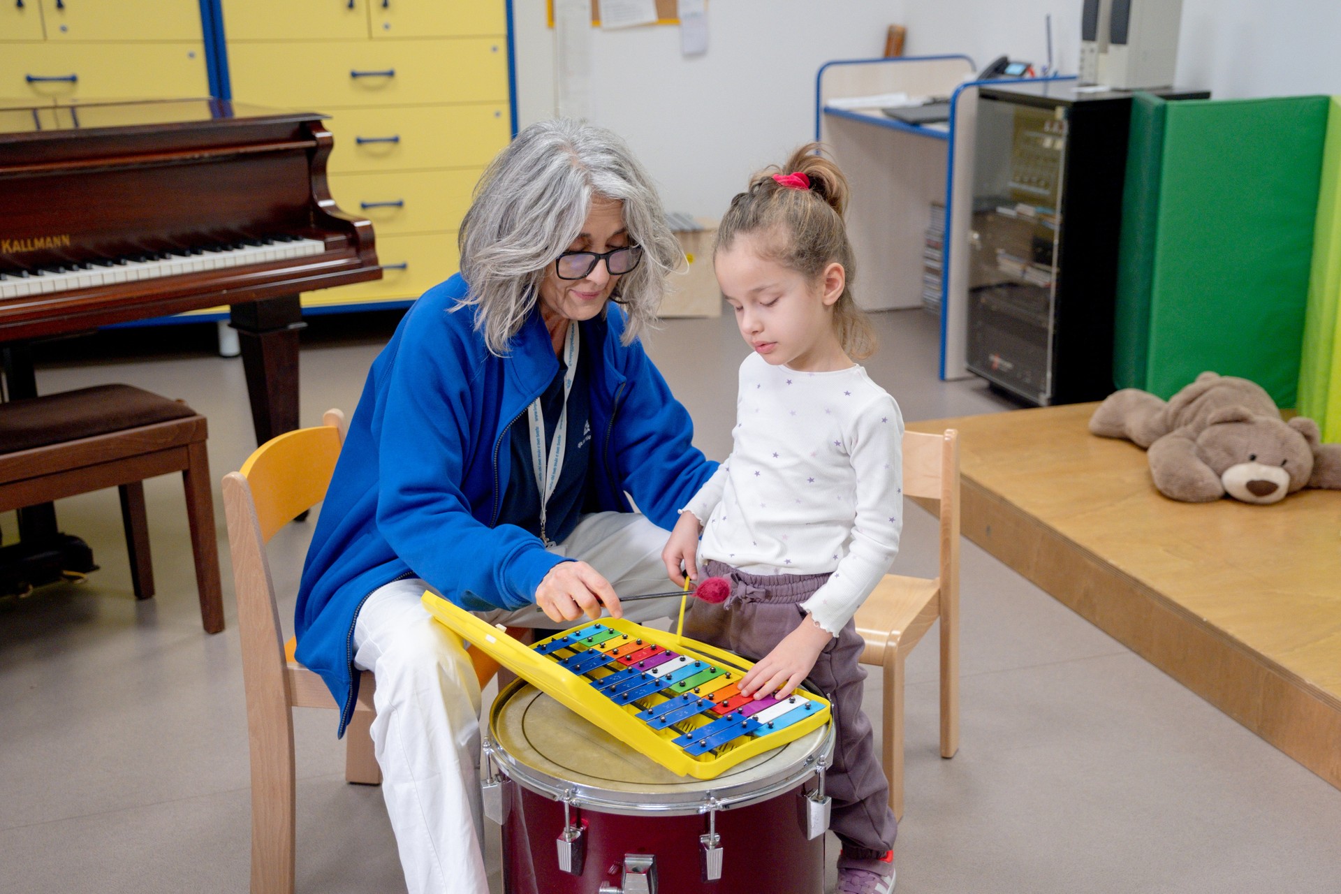 Chiara è in piedi e suona lo xilofono insieme alla sua educatrice, che è seduta a fianco a lei