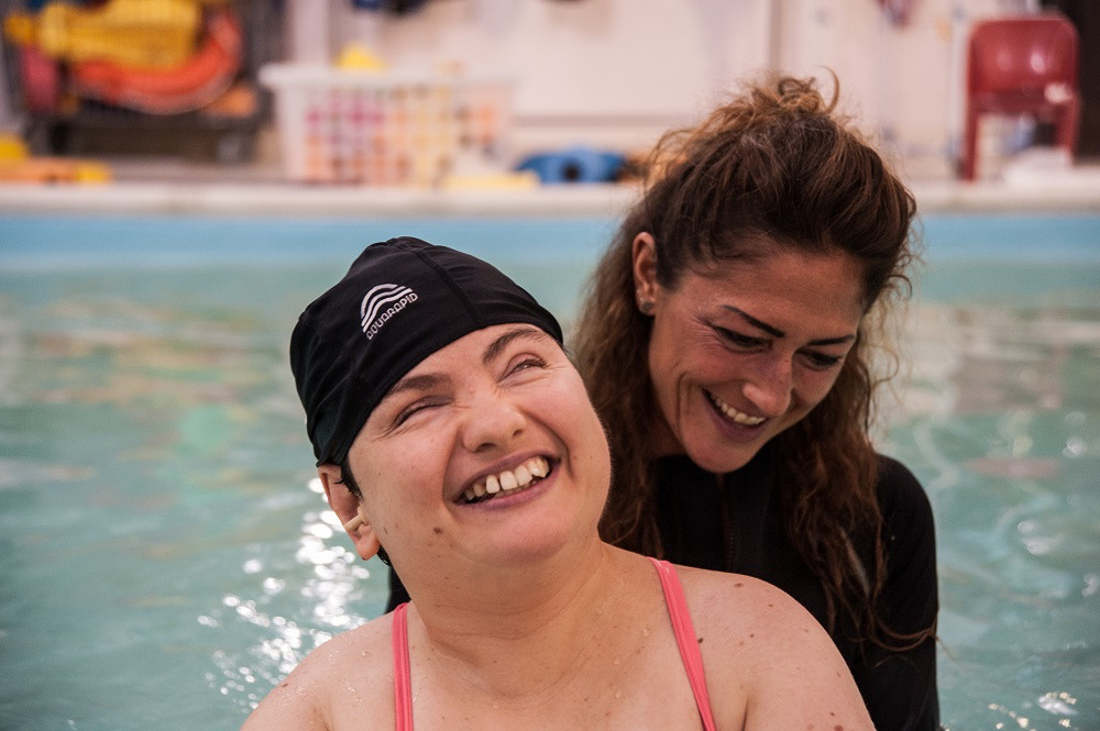 Paola Rupili, utente adulta del Centro di Osimo, sorride insieme alla fisioterapista che la affianca in una sessione di idroterapia in piscina