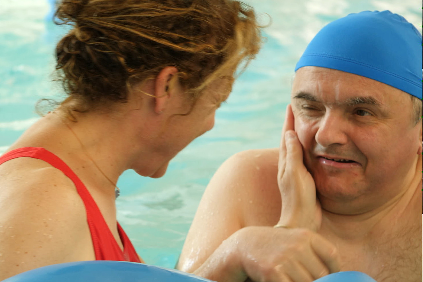 Fabio, utente adulto del Centro di Osimo, è in piscina per fare esercizi. La fisioterapista è di fianco a lui di profilo e gli sorride facendogli una carezza sul viso, Fabio a sua volta sorride.