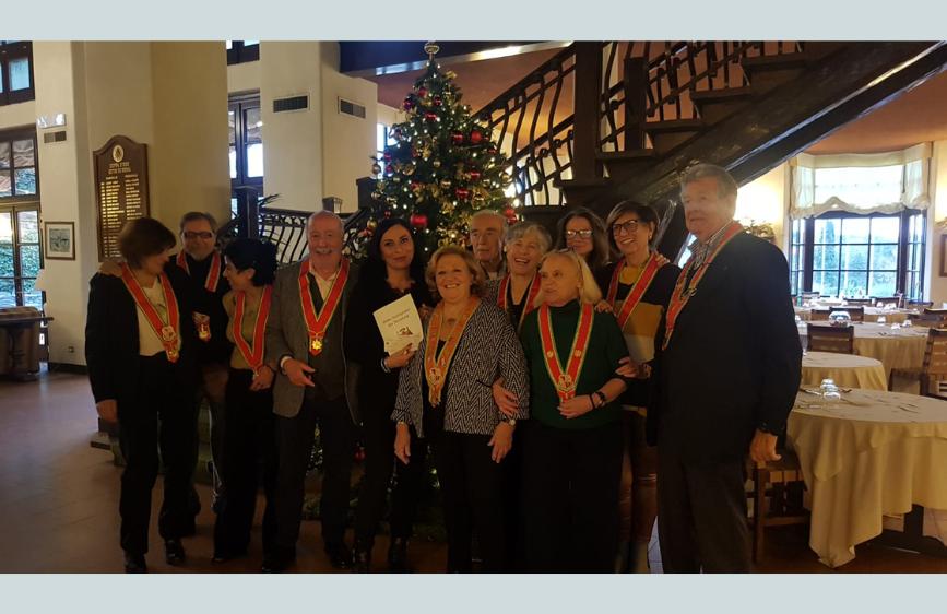 La foto ritrae un gruppo di persone , alcune delle quali indossano collane o fasce cerimoniali rosse e dorate, in posa sorridente davanti a un albero di Natale addobbato, in un ambiente interno che sembra un ristorante o una sala ricevimenti. L'immagine rappresenta un'iniziativa dell'Ordre International des Anysetiers - Commanderie di Roma a favore della Lega del Filo d'Oro.