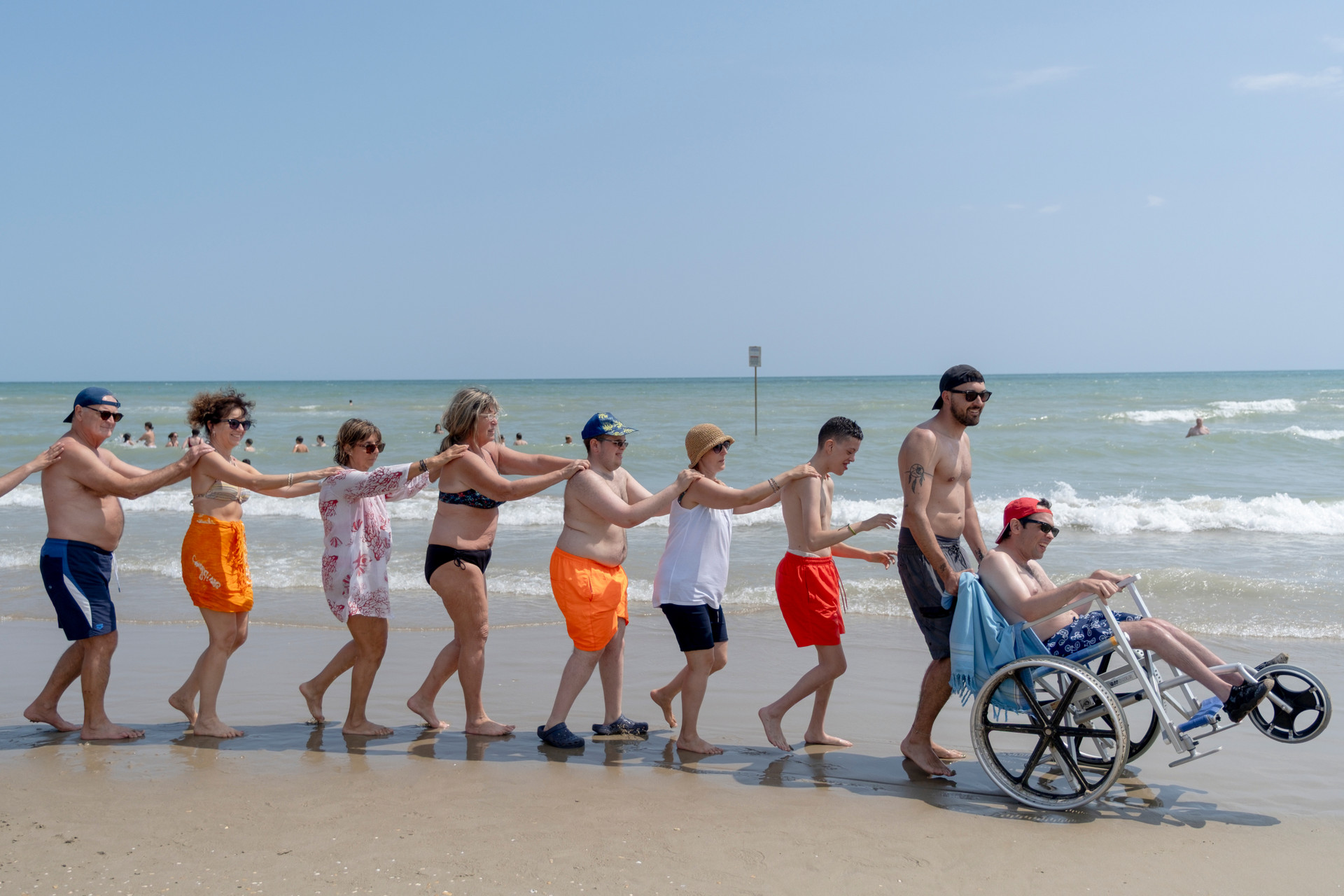 Educatori, ragazzi e ragazze si divertono facendo il trenino sulla spiaggia, accompagnati dalla musica, durante il soggiorno estivo: un momento di festa, inclusione e allegria condivisa.