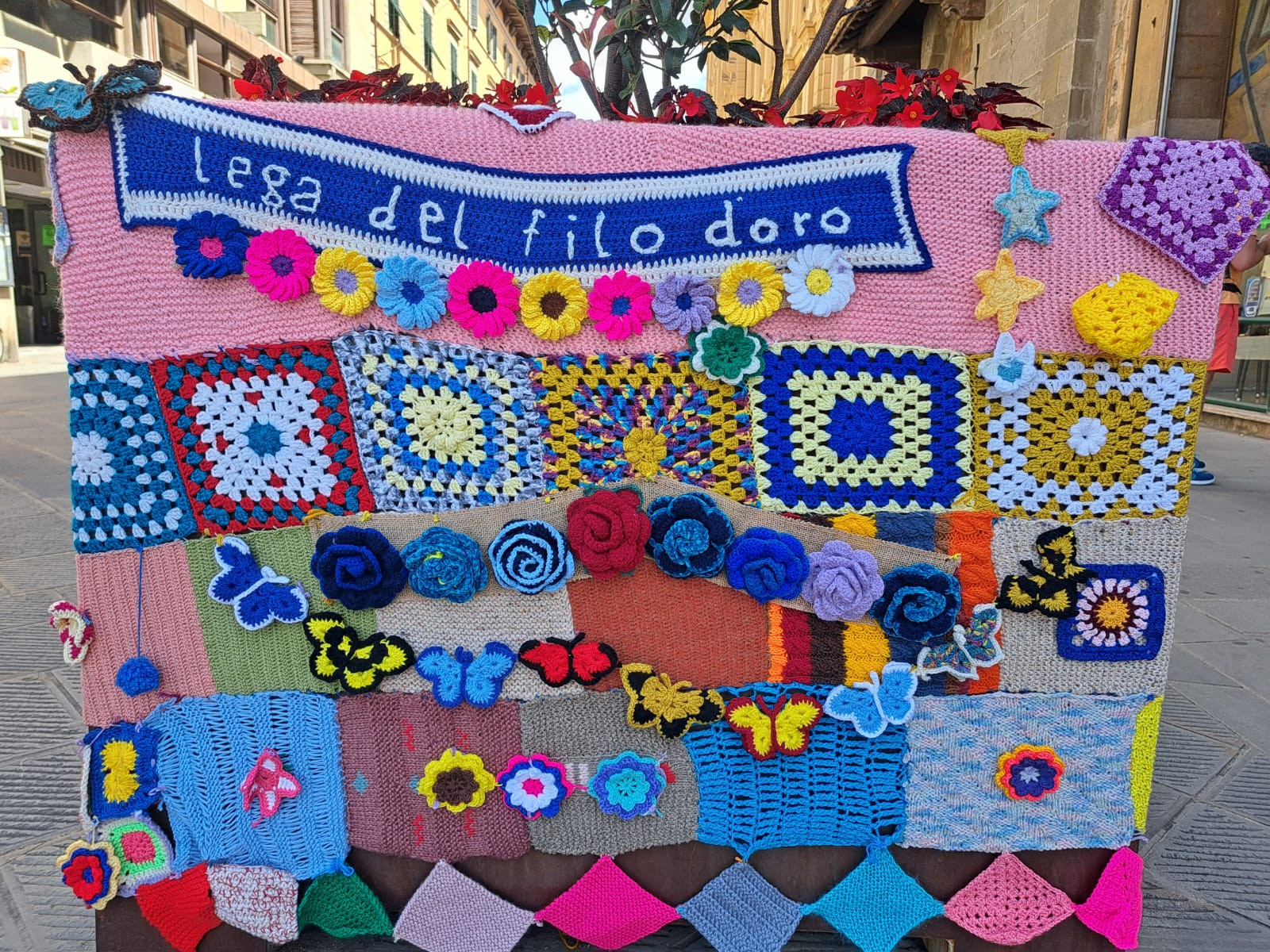 Dettaglio di una coperta fatta a maglia per lo Yarn Bombing a Pisa. Su una sezione blu è visibile la scritta “Lega del Filo d’Oro”, ricamata in bianco.