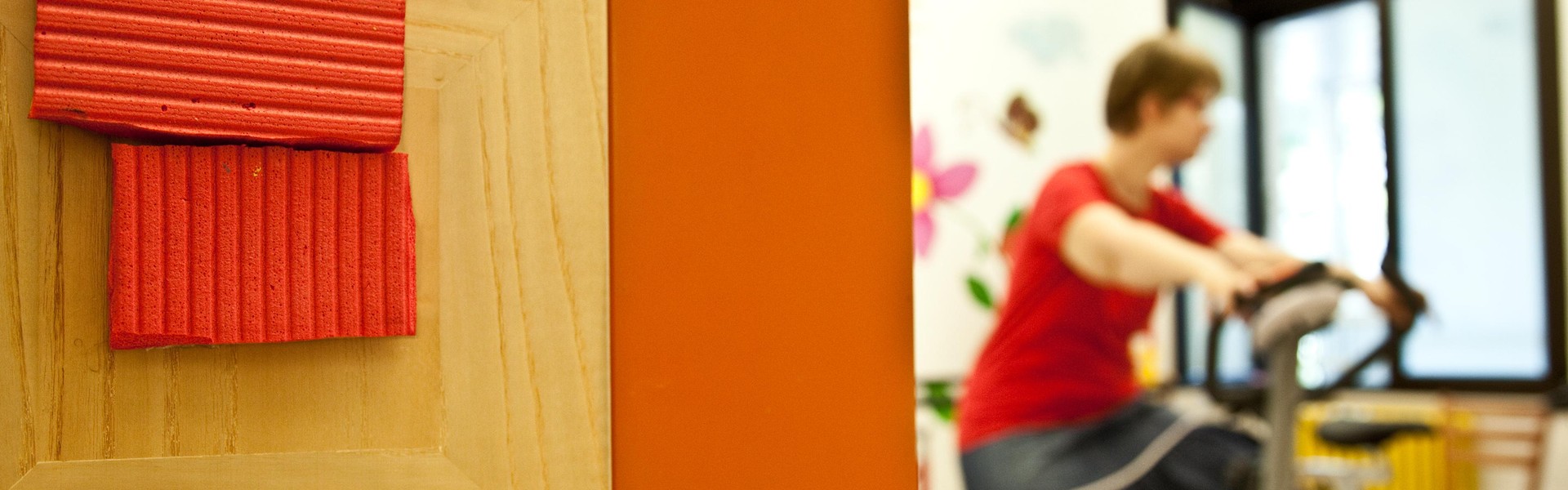 Un segnalino oggettuale con un quadrato di gommapiuma ritagliato da un tappetino per esercizi ginnici, segnala l'ingresso della palestra. All'interno si intravede una utente che fa esercizio sulla cyclette.