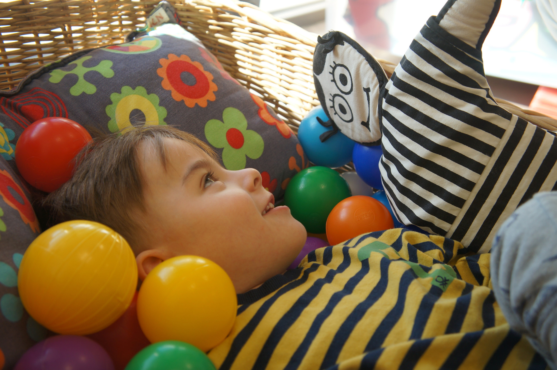 Il piccolo Edoardo, ospite del Centro Diagnostico, sorride immerso in una cesta piena di palline colorate, nel frattempo l'educatrice attira la sua attenzione con un pupazzo di pezza dai forti contrasti cromatici (bianco e nero)