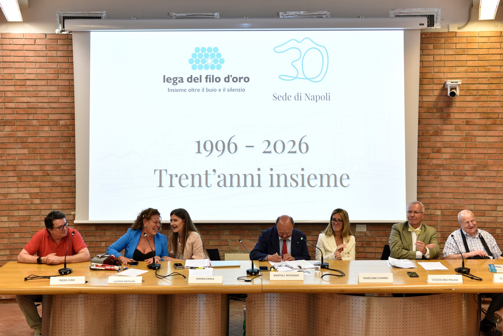 I 6 speaker della presentazione siedono dietro il tavolo al centro della foto. Dietro di loro c'è proiettata l'immagine "1996 - 2026 Trent'anni insieme"