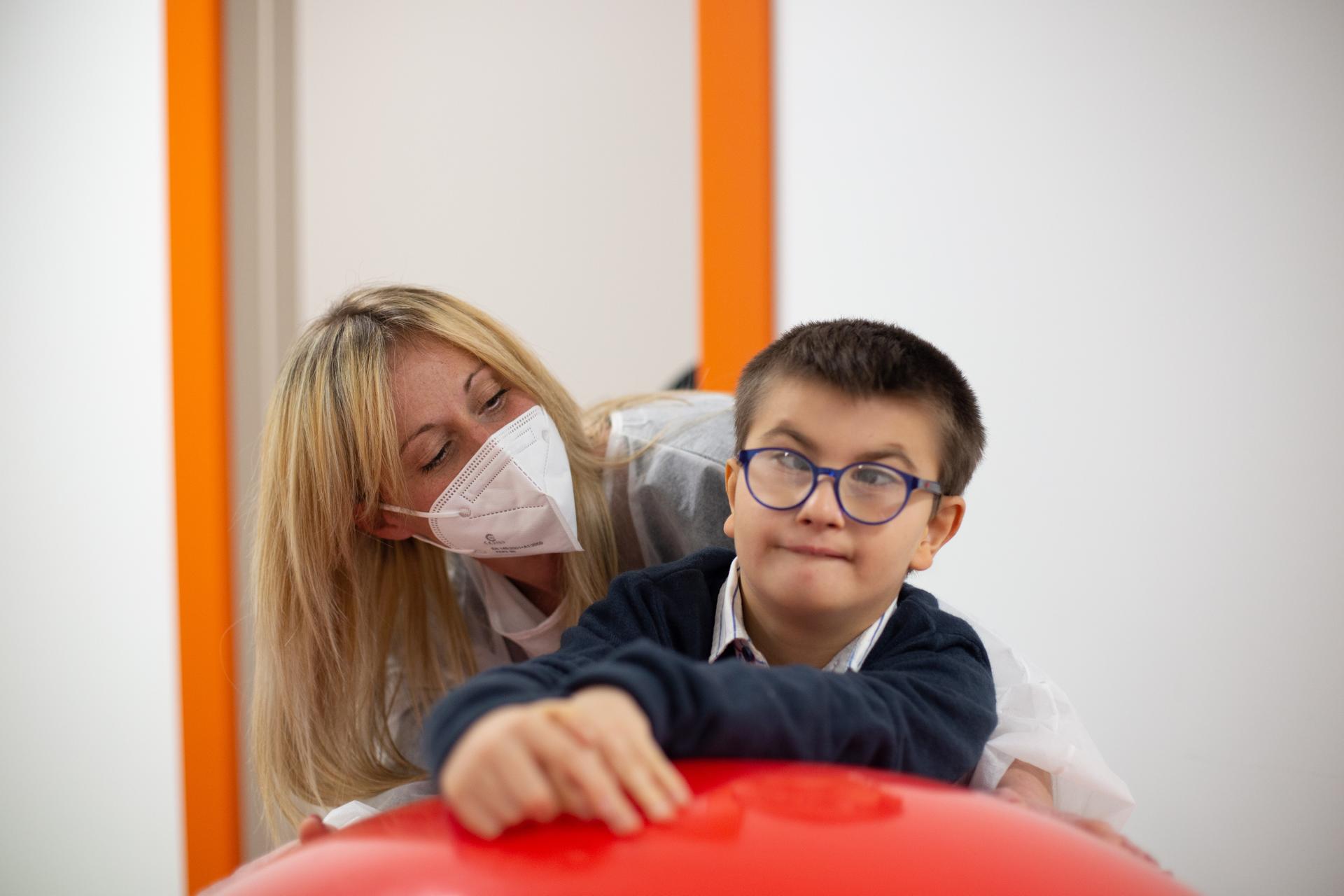 Biagio Luigi, bambino ospite del Centro di Osimo, si appoggia a una grande palla rossa. Alle sue spalle c'è la sua educatrice che indossa mascherina ffp2 e camice monouso anti covid