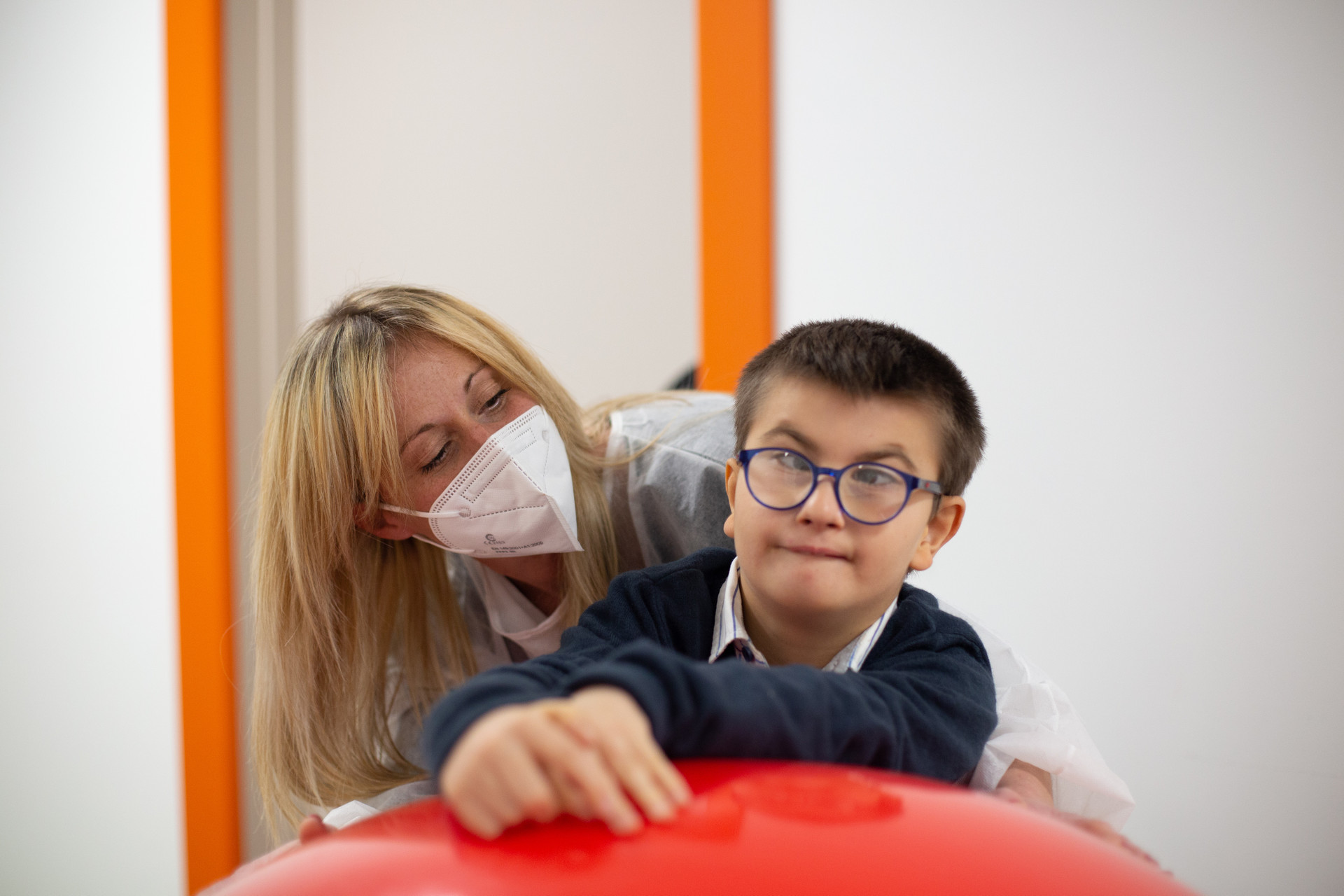 Biagio Luigi, bambino ospite del Centro di Osimo, si appoggia a una grande palla rossa. Alle sue spalle c'è la sua educatrice che indossa mascherina ffp2 e camice monouso anti covid
