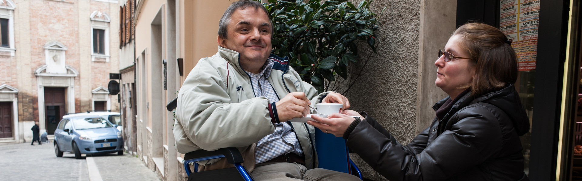 Fabio, utente adulto del Centro di OSimo, è fuori da un bar con la sua carrozzina e sorride bevendo un caffè che gli ha portato la volontaria che lo ha accompagnato