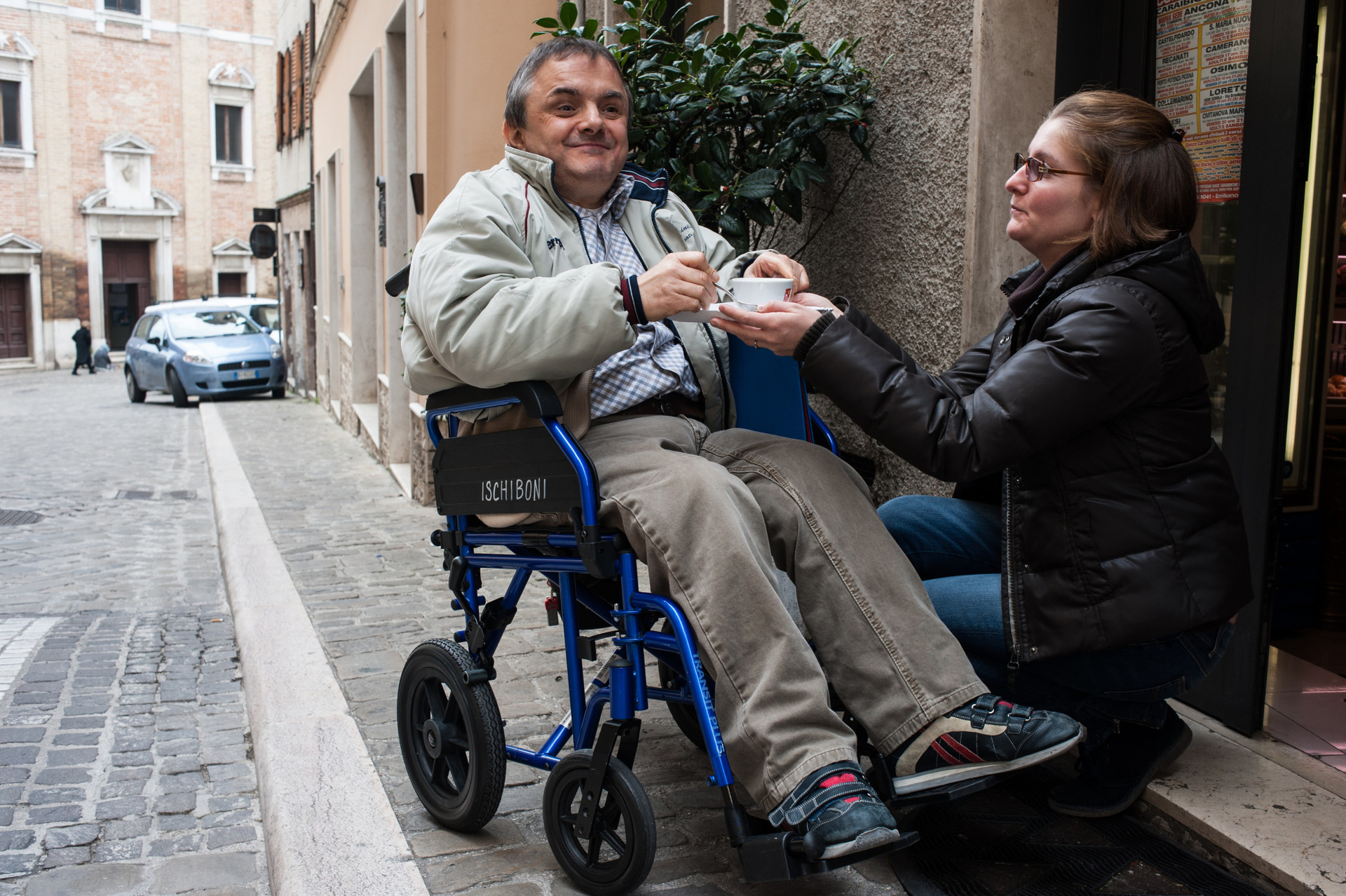 Fabio, utente adulto del Centro di OSimo, è fuori da un bar con la sua carrozzina e sorride bevendo un caffè che gli ha portato la volontaria che lo ha accompagnato