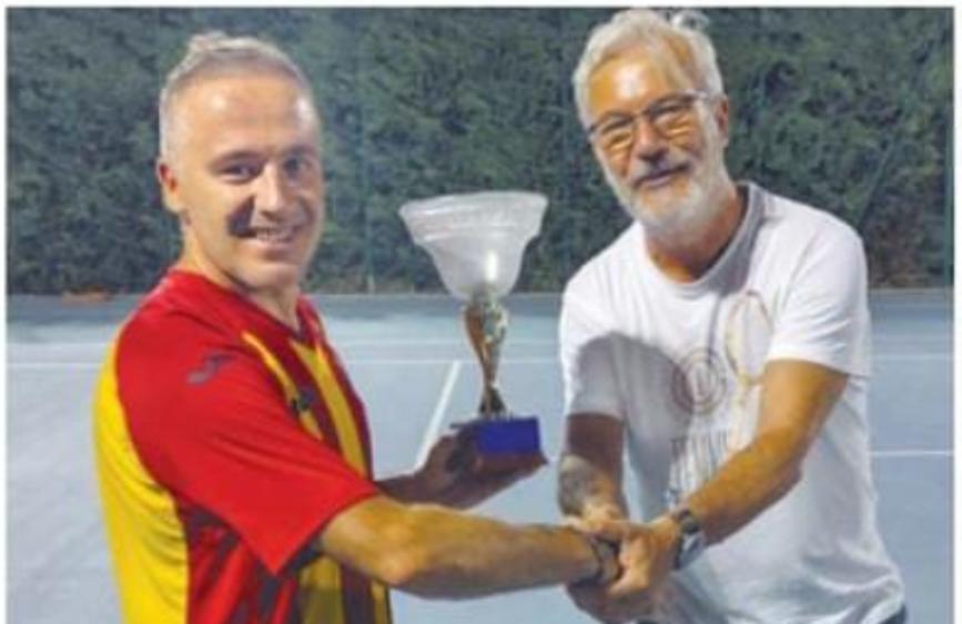 Due uomini si stringono la mano in un momento di premiazione. Uno dei due, in maglia da calcetto rosso-gialla, sorride tenendo una coppa; l'altro, in maglietta bianca e occhiali, gliela sta consegnando. Lo sfondo mostra un campo sportivo illuminato.