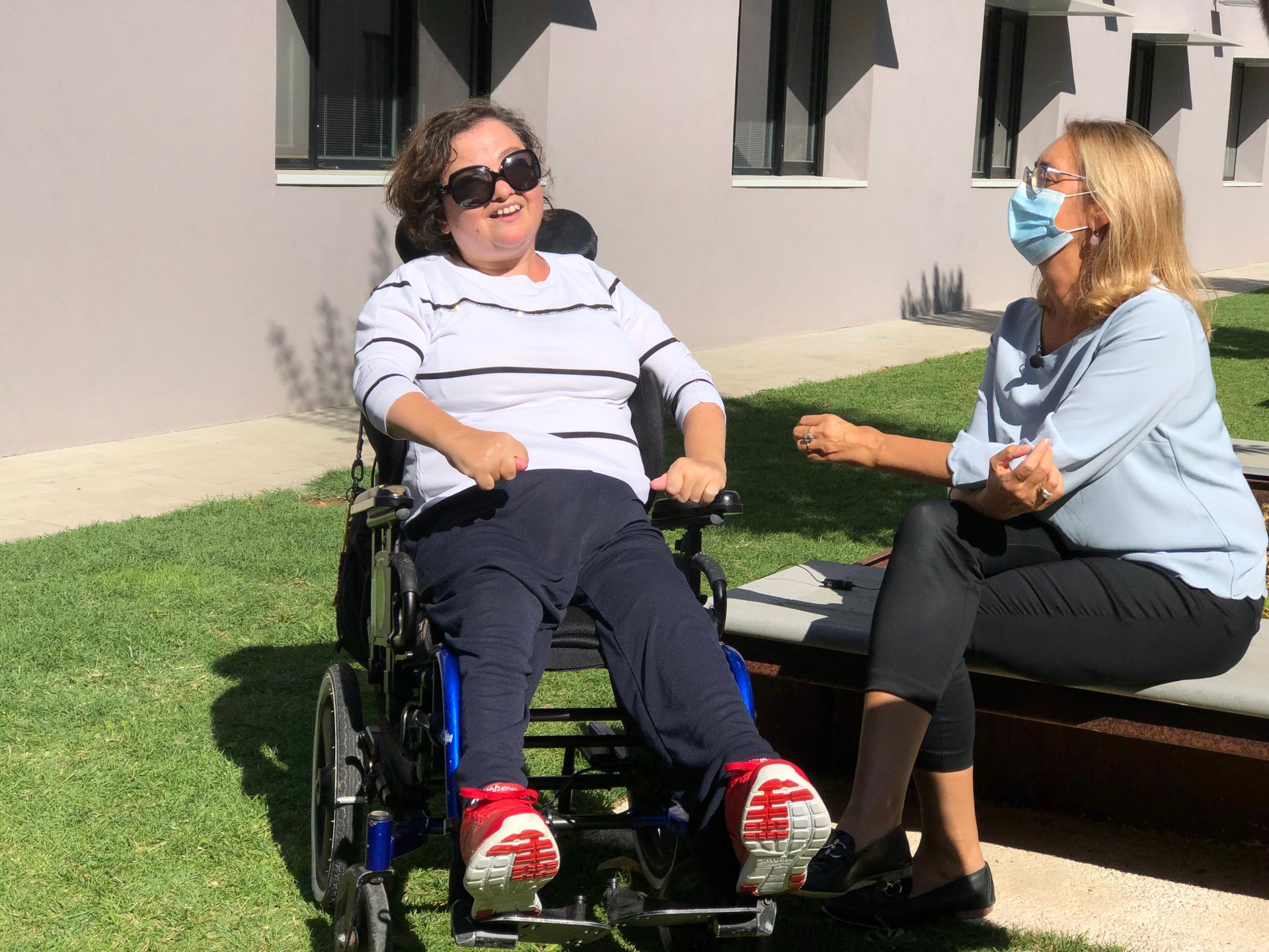 Paola, utente del Centro di Osimo, e Nicoletta si trovano in uno dei giardini del Centro Nazionale. C'è il sole e sorridono mentre parlano tra loro. 