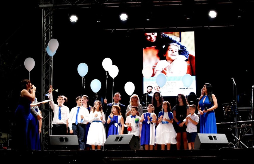 Su un palco illuminato, un gruppo di adulti e bambini in abiti eleganti, molti in blu e bianco, tengono palloncini bianchi e azzurri. Sullo sfondo, un grande schermo mostra la foto di una bambina sorridente con una donna in un momento affettuoso.