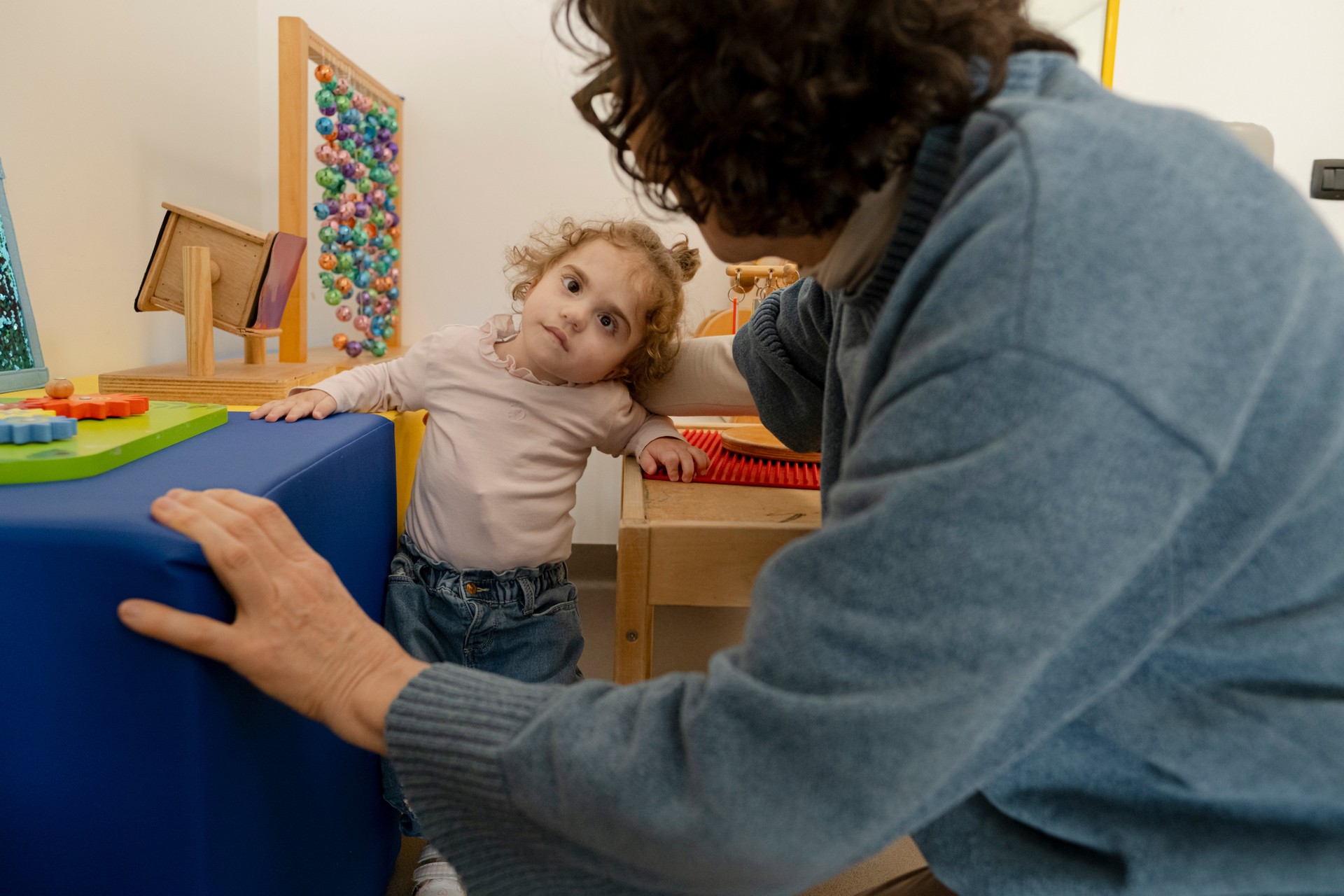 Benedetta, bambina con sindrome CHARGE seguita nel Centro di Osimo, si esercita a stare in piedi con il sostegno della sua educatrice. Intorno a lei ci sono tanti strumenti colorati per la stimolazione sensoriale