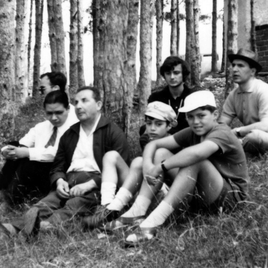 Un gruppo di uomini, alcuni molto giovani, tra cui alcuni utenti della Lega del FIlo d'Oro, si riposano seduti sull'erba in mezzo a un boschetto.