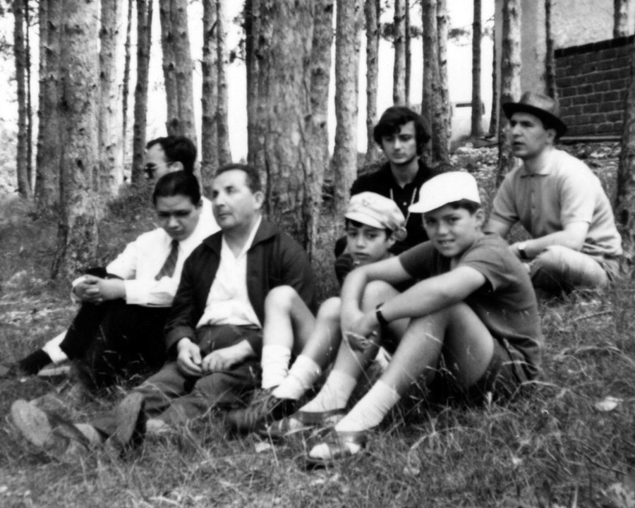 Un gruppo di uomini, alcuni molto giovani, tra cui alcuni utenti della Lega del FIlo d'Oro, si riposano seduti sull'erba in mezzo a un boschetto.