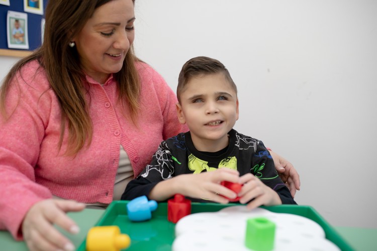 Matteo, bambino con pluridisabilità, è seduto a un banco con la sua educatrice e riconosce al tatto le forme di alcuni oggetti
