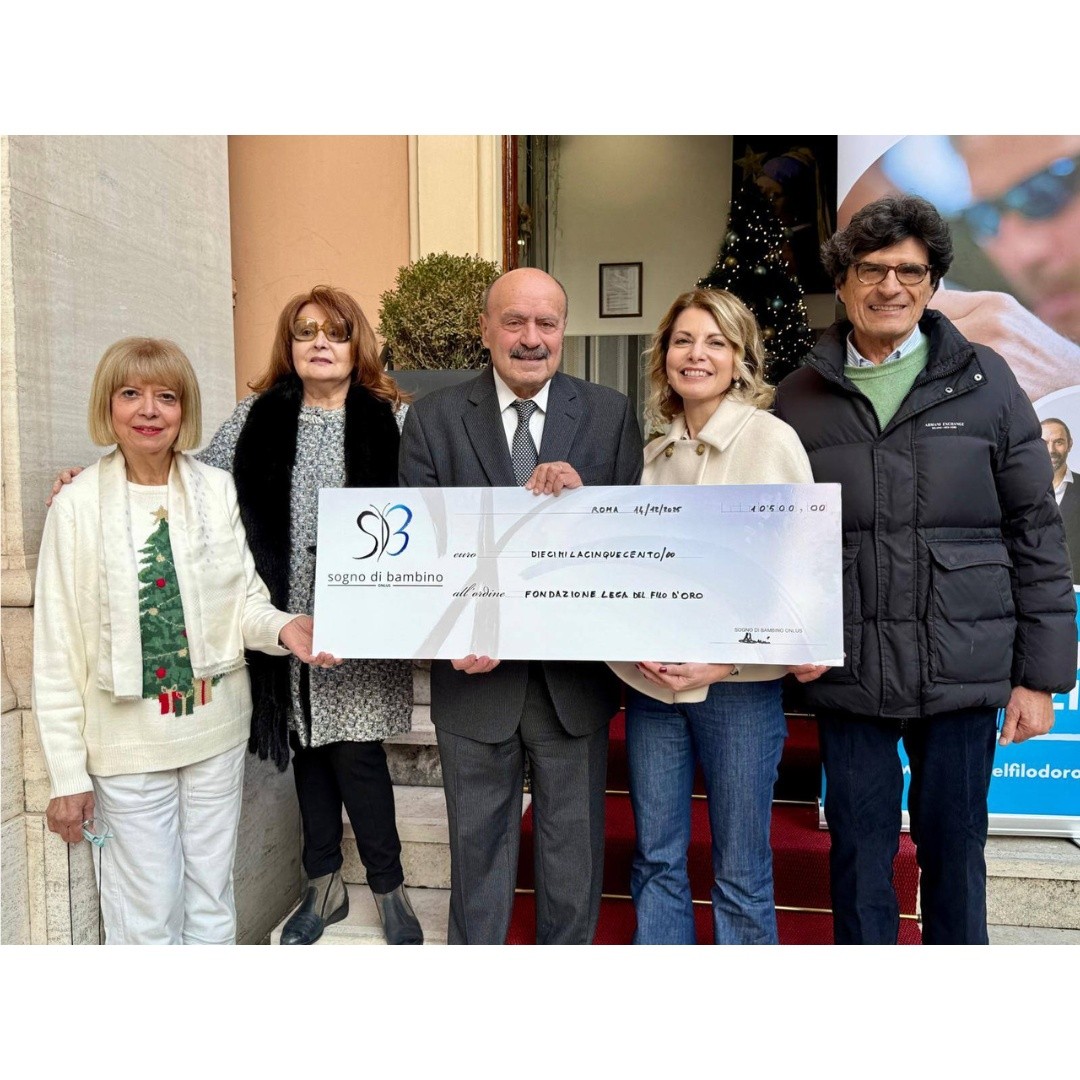 La foto mostra un gruppo di 5 persone in piedi su una scalinata all’ingresso di un edificio. L’ingresso ha una porta aperta e un tappeto rosso che copre i gradini. Sul lato destro, accanto alla porta, c’è un grande banner verticale con il logo e il testo “Lega del Filo d’Oro”. Si tratta della consegan dell'assegno di donazione da Sogno di Bambino a Lega del Filo d'Oro
