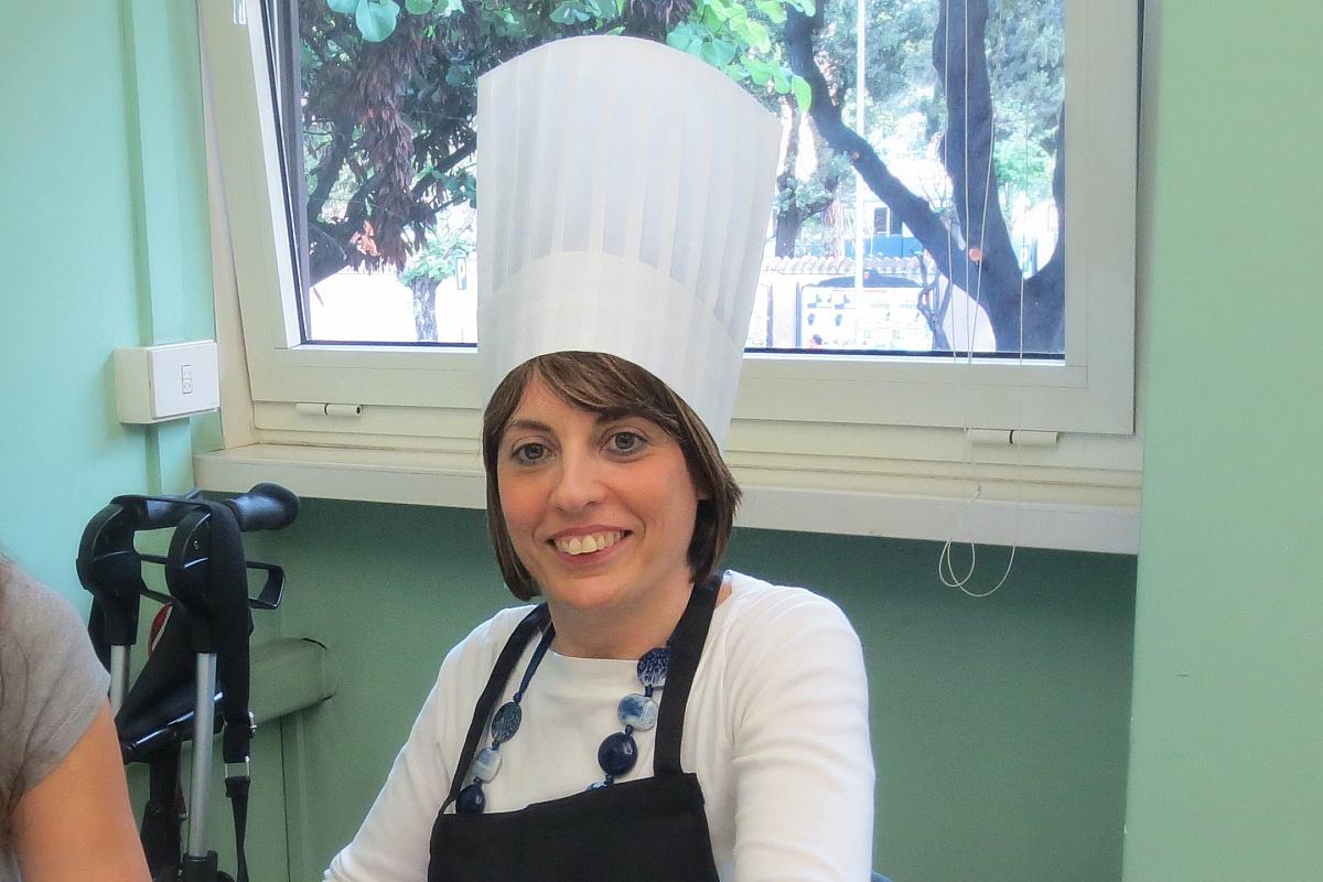 Simona, utente seguita dalla Lega del Filo d'Oro, è ritratta con un grande cappello bianco da cuoco. Completa l'abbigliamento anche un grembiule nero. Sullo sfondo si vede una finestra con vista su un giardino.