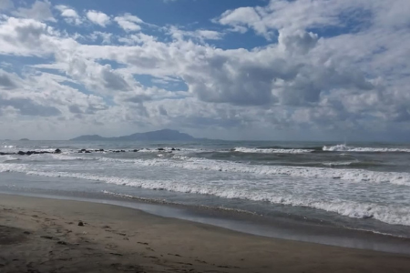 Una spiaggia con il mare mosso e sullo sfondo alcuni pezzi di terra o isole. Le onde del mare fanno schiuma arrivando a riva. Il cielo è nuovoloso.