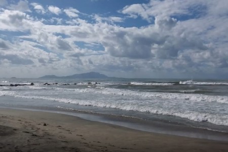 Una spiaggia con il mare mosso e sullo sfondo alcuni pezzi di terra o isole. Le onde del mare fanno schiuma arrivando a riva. Il cielo è nuovoloso. 