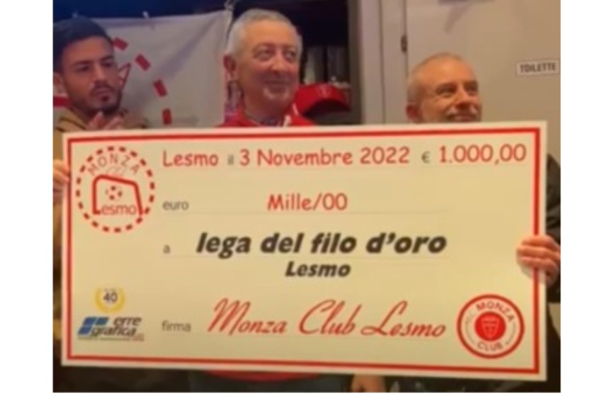 La foto mostra tre uomini, due dei quali reggono un grande assegno simbolico da 1.000,00 euro destinato alla "Lega del Filo d'Oro" da parte del "Monza Club Lesmo". Il contesto suggerisce una cerimonia di donazione o beneficenza.