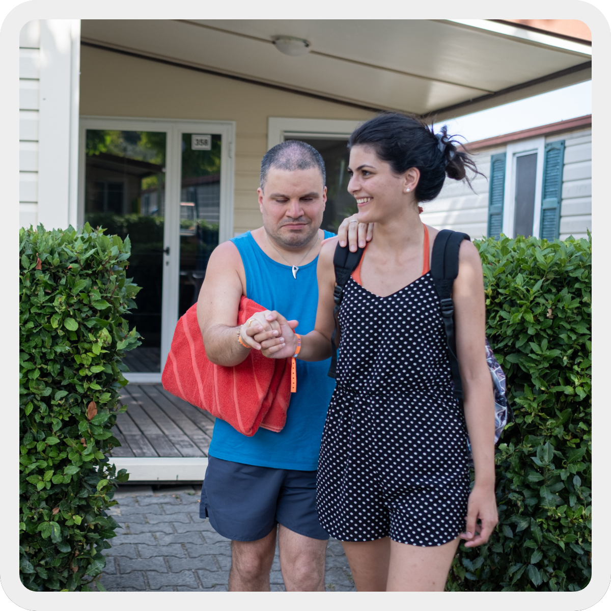 Durante i soggiorni estivi una giovane volontaria accompagna un ragazzo con sordocecità dal suo bungalow alla spiaggia. La volontaria indossa un vestito a pois blu e bianco, il ragazzo una canotta e un paio di pantaloncini blu ed ha un telo mare rosso sottobraccio.