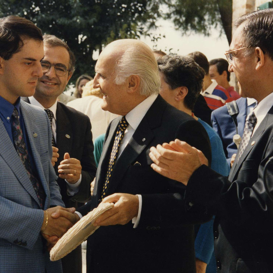 In mezzo alla folla durante una visita alla Lega del Filo d'Oro, il Presidente della Repubblica Oscar Luigi Scalfaro stringe la mano a un utente sordocieco. I presenti assistono alla scena e tutti sono felici.