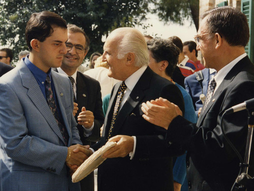 In mezzo alla folla durante una visita alla Lega del Filo d'Oro, il Presidente della Repubblica Oscar Luigi Scalfaro stringe la mano a un utente sordocieco. I presenti assistono alla scena e tutti sono felici.