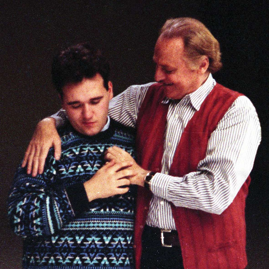 Renzo Arbore e un utente della Lega del Filo d'Oro si abbracciano sul set di una campagna istituzionale. Dietro lo sfondo è neutro.