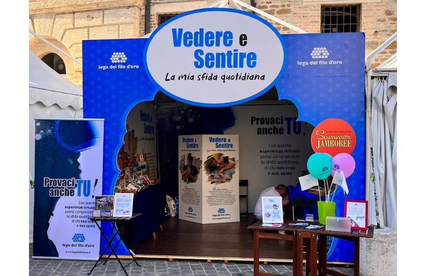 L'immagine mostra lo stand della Lega del Filo d'Oro al Summer Jamboree. Il tema dello stand è "Vedere e Sentire - La mia sfida quotidiana". Lo stand, prevalentemente blu, presenta pannelli informativi e materiale promozionale. A sinistra, un rollup invita il pubblico a "Provaci anche TU!". Sul tavolo di destra ci sono palloncini del Summer Jamboree