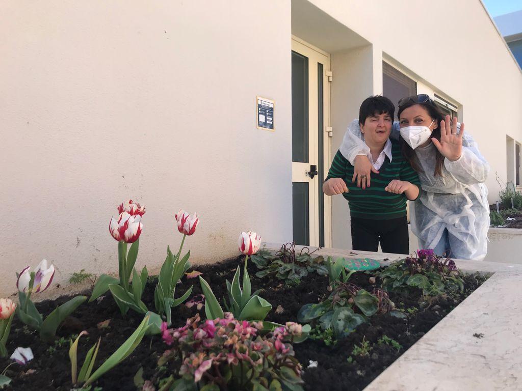 Un utente sorride mentre un'operatrice, vestita con camice e mascherina nel rispetto delle norme anti covid-19, lo abbraccia e saluta verso la macchina fotografica. Davanti a loro, in primo piano, c'è l'orto coltivato dagli utenti del Centro di Termini Imerese, con alcuni fiori rossi e bianchi e altre piantine.
