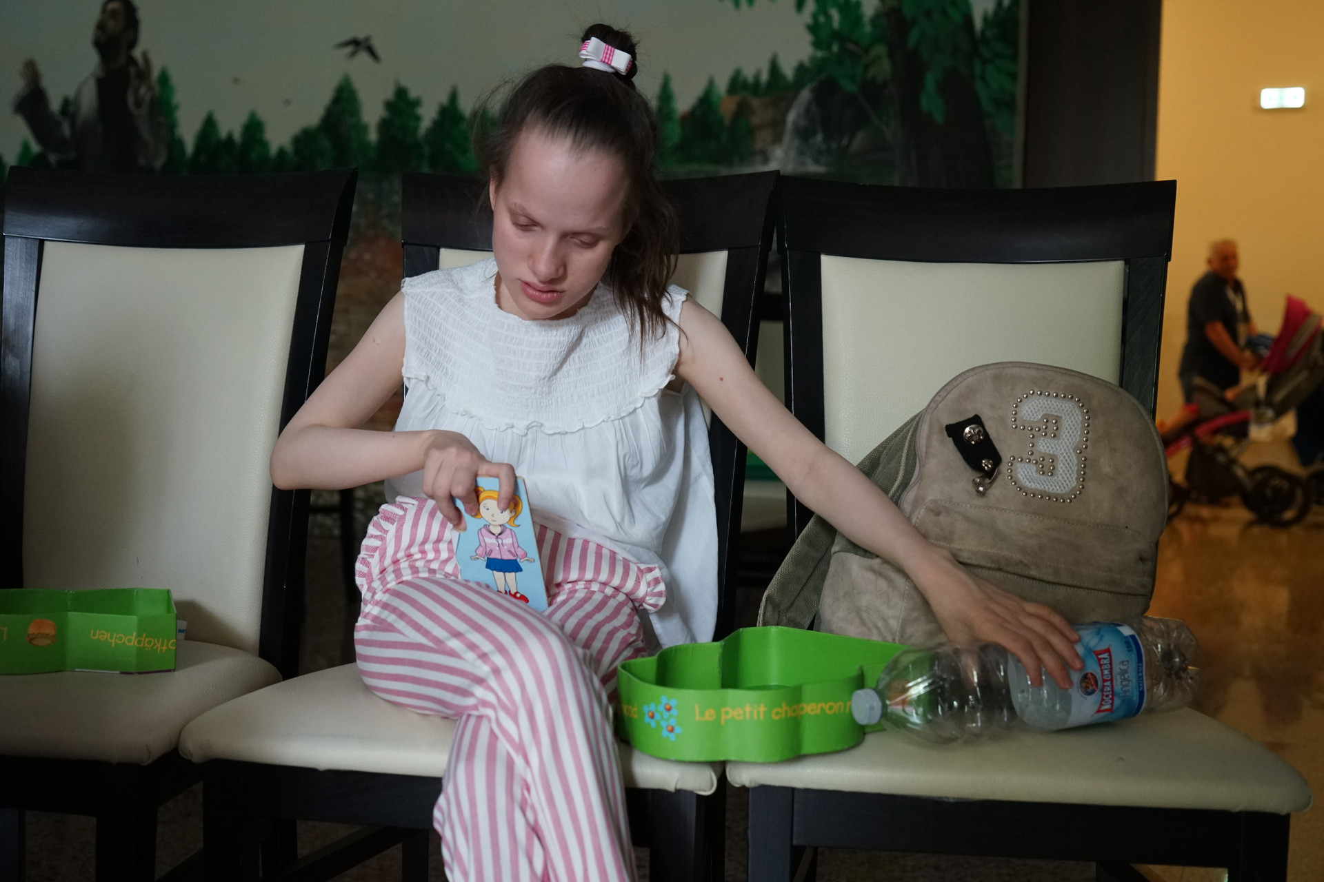  Ragazza sordocieca seduta in una stanza laboratorio, tiene in mano un cartoncino con un disegno e una bottiglia di plastica. Lo sguardo è rivolto verso il basso.