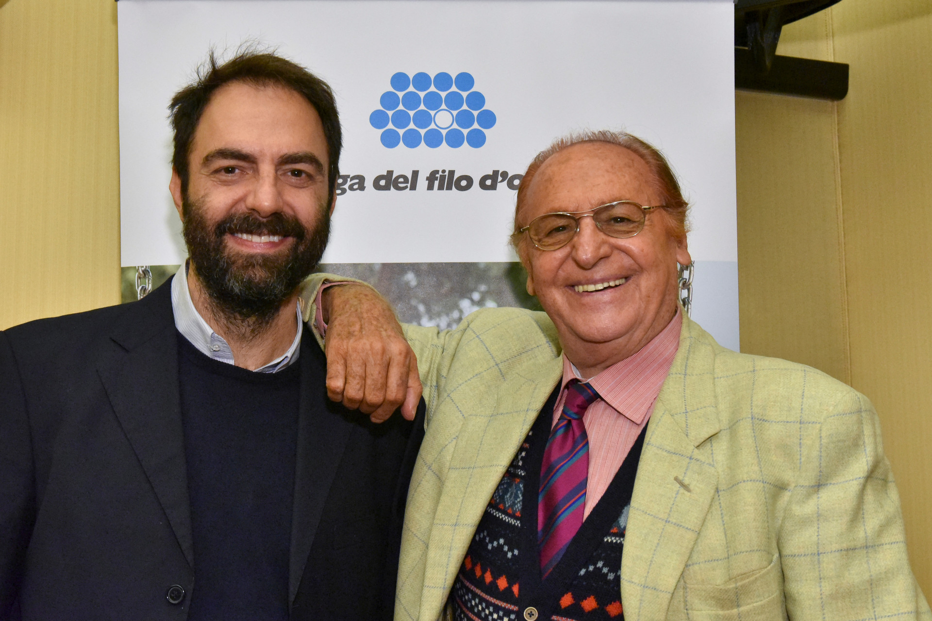 Renzo Arbore e Neri Marcore sorridenti posano per una foto di fronte al logo Lega del Filo d'Oro