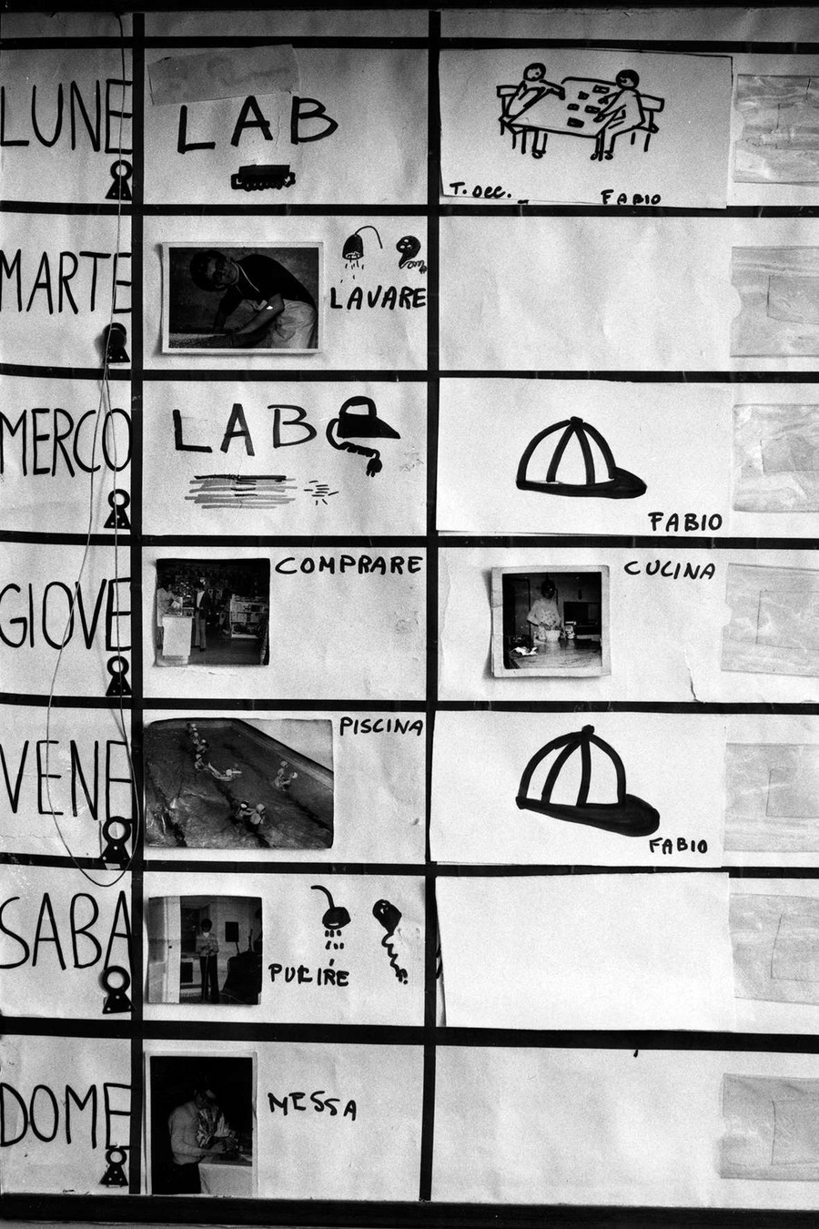 In bianco e nero, la foto ritrae un calendario pittografico, con una tabella in cui sono segnati i giorni della settimana, le attività e gli incaricati di queste.