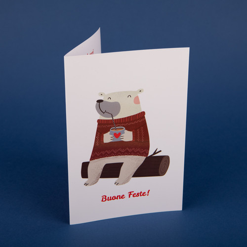 Biglietto di auguri in cartoncino, con Disegno su sfondo bianco di un orsacchiotto bianco sorridente, seduto su un tronco d'albero e beve una cioccolata calda da una tazza decorata con un cuoricino. Sotto il disegno la scritta "Buone Feste!" in rosso. 6