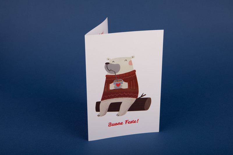 Biglietto di auguri in cartoncino, con Disegno su sfondo bianco di un orsacchiotto bianco sorridente, seduto su un tronco d'albero e beve una cioccolata calda da una tazza decorata con un cuoricino. Sotto il disegno la scritta "Buone Feste!" in rosso.