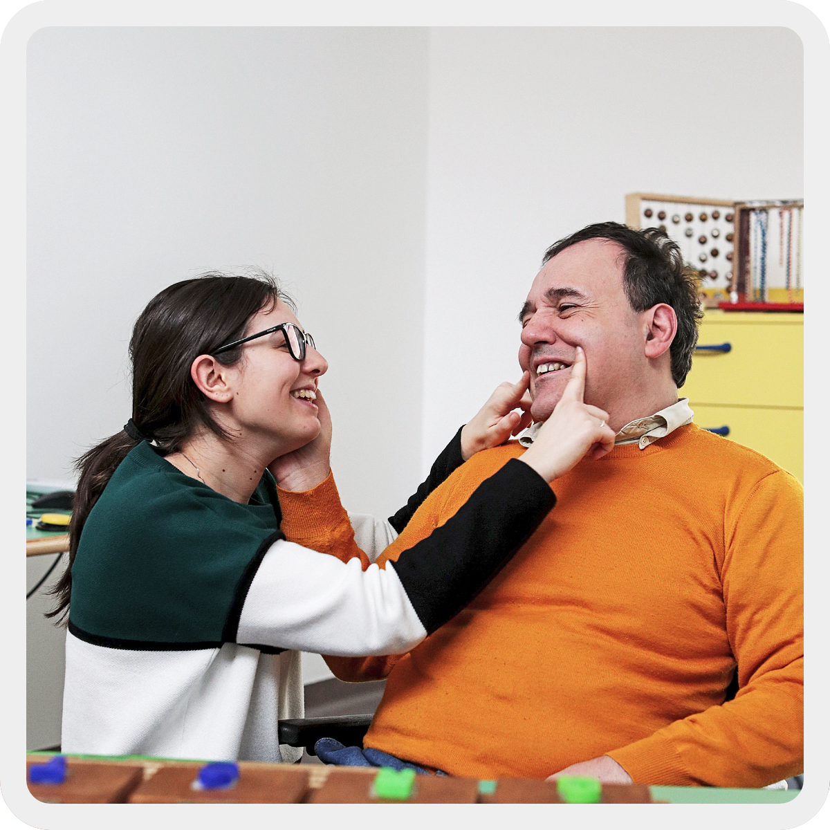 Alberto, adulto con pluridisabilità sorride e accarezza il viso di Micol, la sua educatrice, che sorridendo anch'essa tiene le due dita indice ai lati della bocca di Alberto per evidenziare ancora di più il suo sorriso. Sono all'interno di un'aula del Centro Nazionale. 
