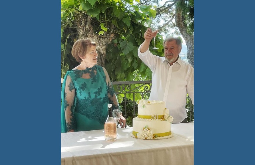 L'immagine celebra il 50° anniversario dei coniugi Poeta. La foto ritrae un uomo con una camicia bianca che alza un calice in un brindisi, mentre sua moglie, indossa un elegante abito verde smeraldo e guarda verso di lui. Di fronte a loro c'è una torta nuziale a due piani decorata con rose bianche e il numero 50 in oro, il tutto su un tavolo imbandito all'aperto, con una lussureggiante vegetazione sullo sfondo.